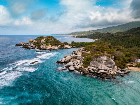 Aerial view of the rocky coastline and turquoise waters of Parque Nacional Natural Tayrona, featuring large granite boulders, lush tropical vegetation, and a sandy beach in Magdalena, Colombia.