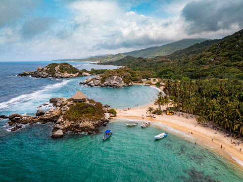 Aerial view of Parque Nacional Natural Tayrona featuring a sandy beach, turquoise water, rocky outcrops, palm trees, and boats under a cloudy sky in Magdalena, Colombia.