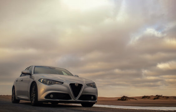 Silver Alfa Romeo Giulia sedan parked on the coast under a dramatic cloudy sky at sunset. Luxury Italian automobile vehicle on a road trip. Elegant transport in a scenic nature landscape background. 