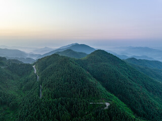 Fototapeta premium Layered forested mountain ranges with winding roads, shrouded in soft morning mist under a pale dawn sky.