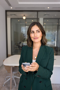 Confident Hispanic businesswoman in elegant jacket holding smartphone and looking aside thoughtfully stands in modern office boardroom. Decision-making, strategic thinking, tech in business, planning