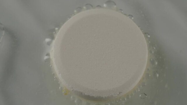 Extreme close-up of a white effervescent tablet dissolving in a glass of water, releasing bubbles and fizzing as it dissolves for pain relief, showcasing a chemical reaction in slow motion
