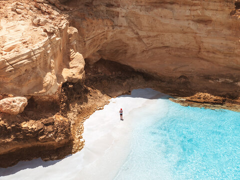 Aerial view of Shoab Beach with turquoise water, white sand, and rocky cliffs, with a person standing on the beach, Qalansiyah, Yemen.