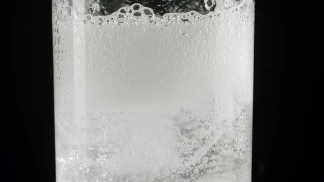 Clear fizzy drink with ice cubes inside a glass, showing bubbles rising slowly to the surface. Fizzy sparkling water with ice cubes and bubbles. White dissolving pill creating fizzy bubbles