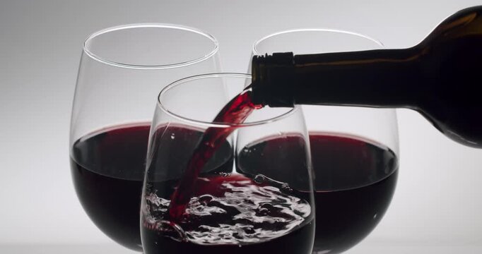 Luxurious red wine pouring from a dark glass bottle into an elegant stemless wineglass in slow motion, isolated on a white background, creating swirls and bubbles for a tasting event