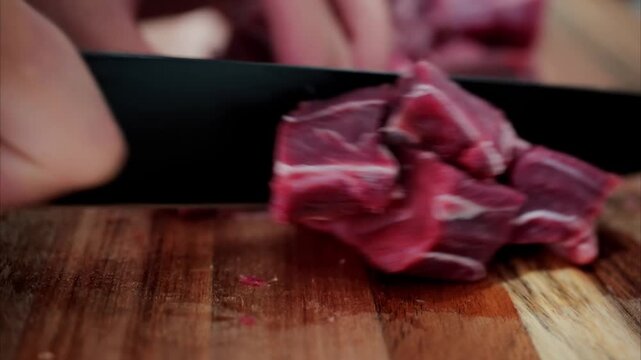 Close up of person cutting raw beef meat into cubes on wooden board for cooking.