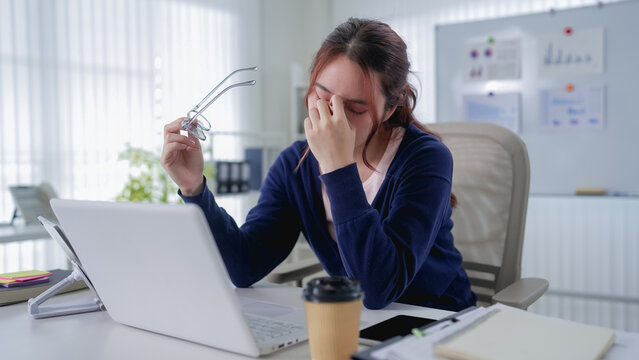 Exhausted young Asian businesswoman rubbing eyes while working at laptop in bright home office with copy space
