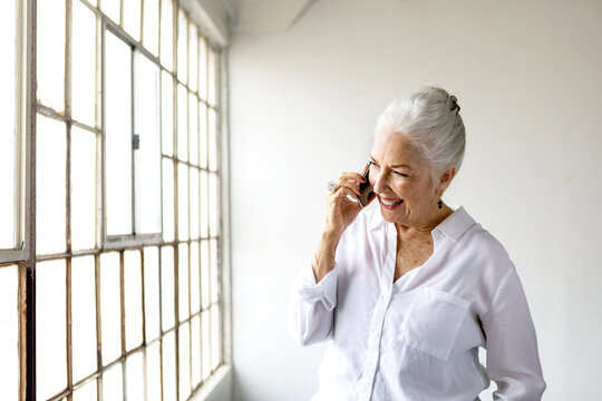 Elderly woman with white hair smiling while talking on the phone by a window.
