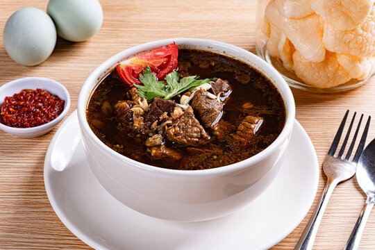 Indonesian rawon black beef soup served with salted eggs, sambal, and crackers