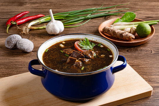 Indonesian rawon dark soup in enamel pot with garlic and green onions on wooden table
