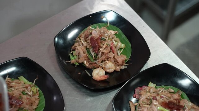 A chef is arranging freshly cooked flat rice noodles&mdash;complete with vegetables and meat&mdash;on a banana leaf, showcasing the process of preparing an Asian dish in a restaurant kitchen.