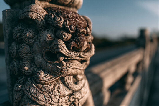 Close-up of weathered ancient stone lion on bridge railing, cinematic golden hour lighting against cool blue sky, shallow depth of field, perfect for cultural heritage and historical documentary use.