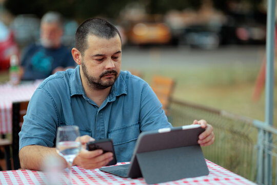 Focused Business Man Using Smartphone And Digital Tablet At Restaurant. Busy adult guy multitasks with mobile devices sitting at outdoor terrace