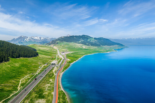 Aerial view of Sayram Lake and mountain landscape in Xinjiang, China, featuring a turquoise shoreline, alpine meadow, forest, and scenic highway.