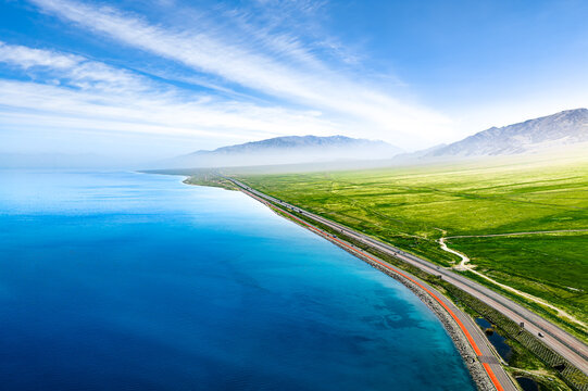 Aerial view of turquoise Sayram Lake shoreline, highway across green grassland beneath the Tien Shan mountains in Xinjiang, China.