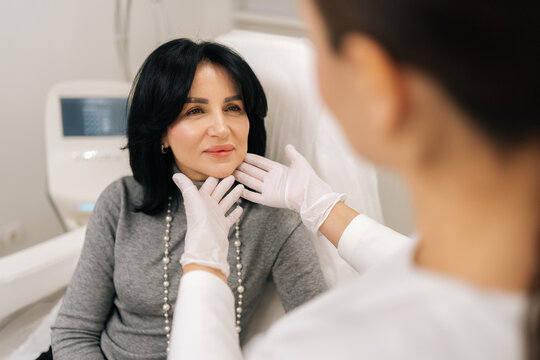 Close-up of professional female cosmetologist wearing gloves talking to mature woman patient in chair and examining facial skin before acosmetic procedure in modern aesthetic clinic.