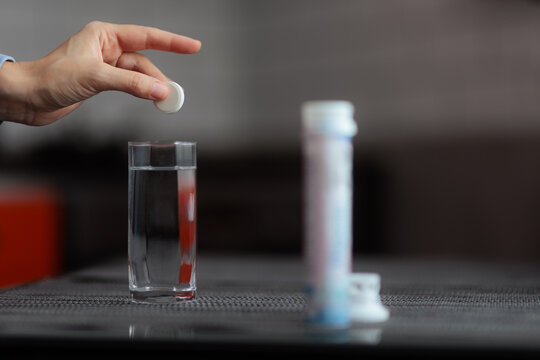 Close Up Of Hand Dropping Effervescent Tablet Into Glass Of Water. Person dissolves a soluble effervescent pill inside a clear glass indoors