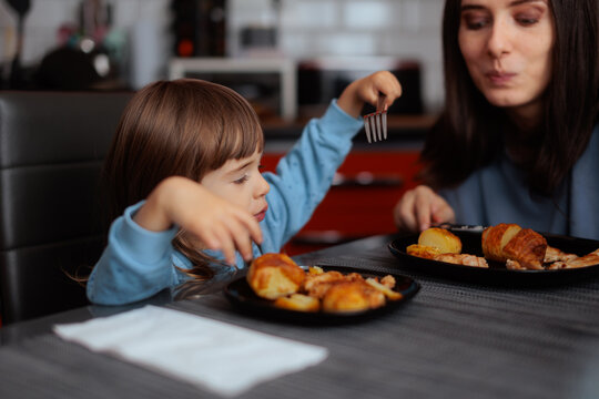 Happy Mother And Little Daughter Having Tasty Dinner At Kitchen Table. A smiling mom looks at her cute child eating food with fork
