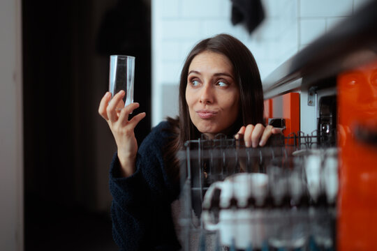 Skeptical Young Woman Unloading Clean Glasses From Dishwasher In Kitchen. Unsure girl inspects a clear glass while doing domestic chores indoors
