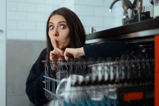 Surprised Young Woman Holding Finger To Lips Near Open Kitchen Dishwasher. Housewife makes a shushing gesture while unloading clean dishes indoors