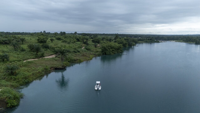 Aerial view of a small boat cruising on the blue waters of McCarthy beach, surrounded by lush green vegetation and palm trees under a cloudy sky in Urhuoka, Delta, Nigeria.