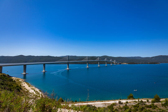 A modern cable-stayed bridge spans across the blue sea connecting landmasses under a clear sky.