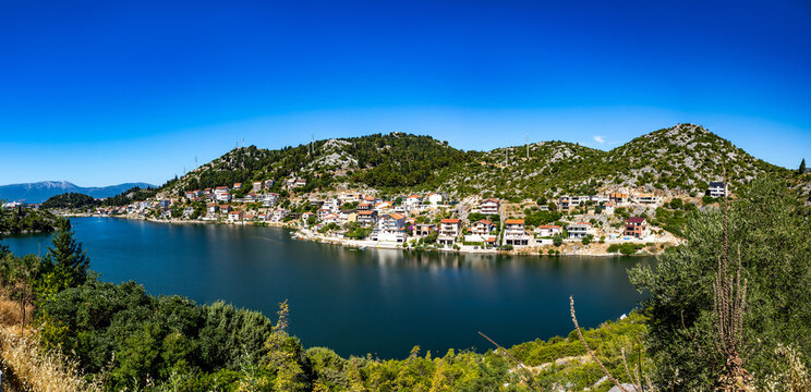A hillside village with Mediterranean-style houses overlooking a calm bay on the Adriatic coast. The landscape features rocky terrain, green vegetation, and clear blue sky.
