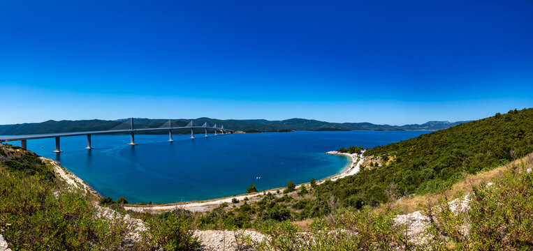 A modern cable-stayed bridge spans across the blue sea connecting landmasses under a clear sky.