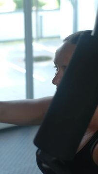 Vertical video. Young woman in black activewear operating a chest press machine in a bright gym space with a focused expression while pressing. Resistance training concept