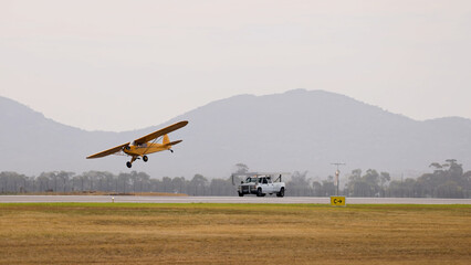 Fototapeta premium Yellow Vintage Piper Cub Aircraft Taking Off Beside a White Truck