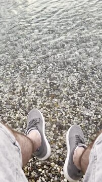 A man in sneakers is sitting on a stone pier above the sea and moves their legs in a relaxed manner above the water. Point of view.