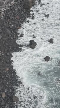 Vista a&eacute;rea cenital desde dron de olas espumosas del mar Atl&aacute;ntico llegando de forma r&iacute;tmica a la orilla de una playa de piedras volc&aacute;nicas en las Islas Canarias