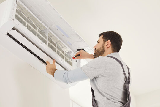 Skilled male mechanic or repairman repairing conditioner at client house on white wall, wearing uniform, assembling air conditioner with screwdriver. Construction and repairing concept