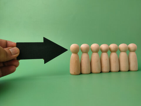 A hand holds a black directional arrow pointing toward a line of seven wooden peg figures on green