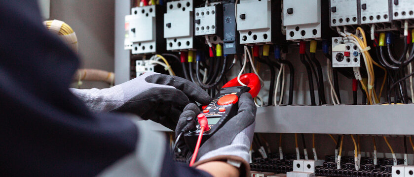 Electrician using digital clamp meter to measure electrical current in control panel.
