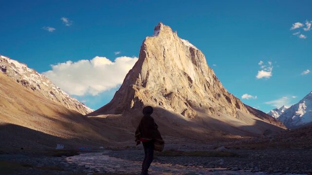 Silhouette shot of an Indian male tourist wearing hat and shawl standing in front of the Holy Gonbo Rangjon mountain during the sunset at Zanskar valley in remote region of Ladakh, India. 