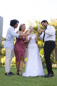 Diverse wedding group on grassy lawn beside wall, bride wearing white gown, groom pouring wine