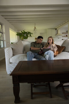 Diverse couple sitting on light-colored sofa in living room using tablet near coffee table