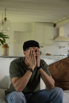 Man sitting on sofa in open-plan living room covering face with hands, wearing wristwatch