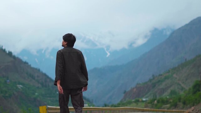 Rear view shot of an Indian male tourist walking in front of Himalayan mountain peaks covered by clouds during the monsoon season near Triloknath in Lahaul and Spiti district, Himachal Pradesh, India.