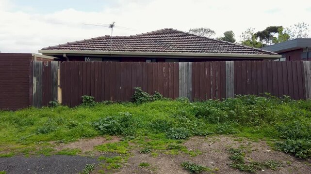 A suburban house behind an aged wooden pailing fence, featuring tiled roofing, an overgrown grassy verge with weeds. Urban neglect and run-down residential neighborhood in Australia.