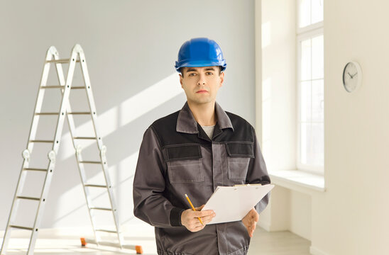 Builder contractor worker foreman handyman inspector renovation safety. Serious male in blue helmet reviews clipboard near window, planning repair. Professional renovation safety concept portrait.