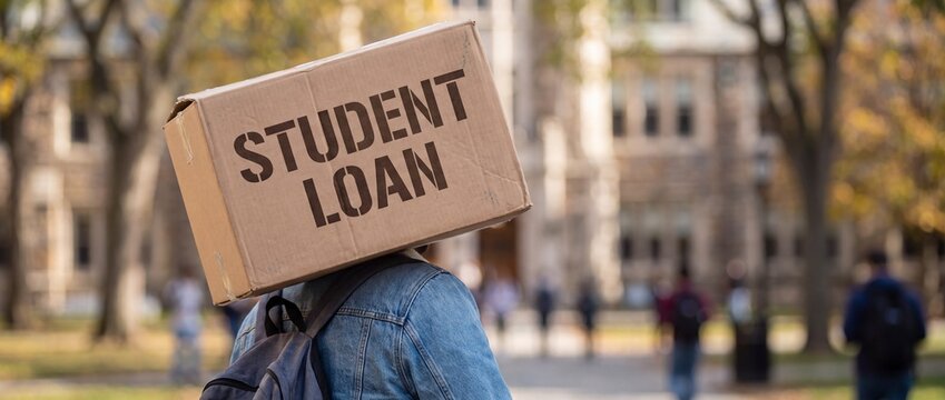 Student carrying a cardboard box labeled student loan on their head in a college campus park during autumn with backpack balancing cardboard
