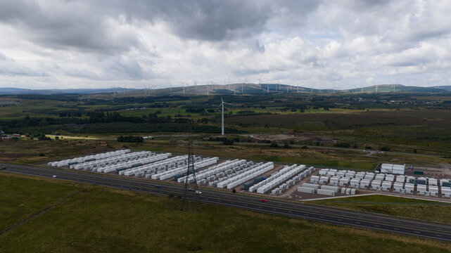 Aerial view of the BESS battery energy storage system facility featuring rows of white containers, a wind turbine, and rolling hills in Lanark, Scotland, United Kingdom.