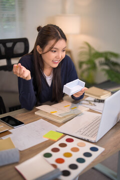 Young Asian furniture designer reviewing color palette and material samples at office desk with laptop