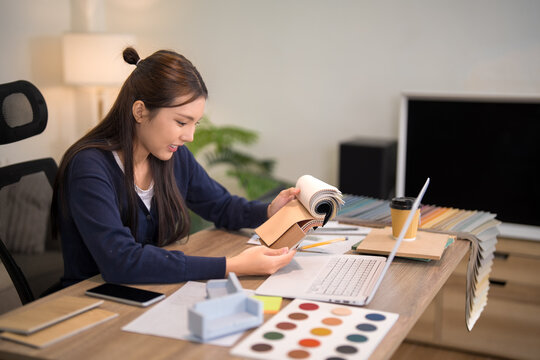 Young Asian furniture designer reviewing material samples and color swatches at wooden desk in modern design studio