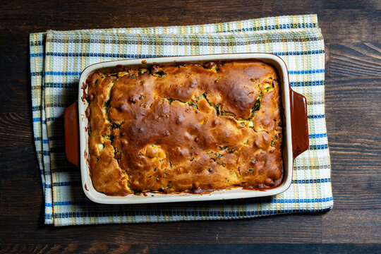 Homemade baked savory pie in ceramic dish. Freshly baked golden brown pie or casserole with herbs and cheese in rectangular ceramic baking pan on checkered towel over rustic dark wooden table