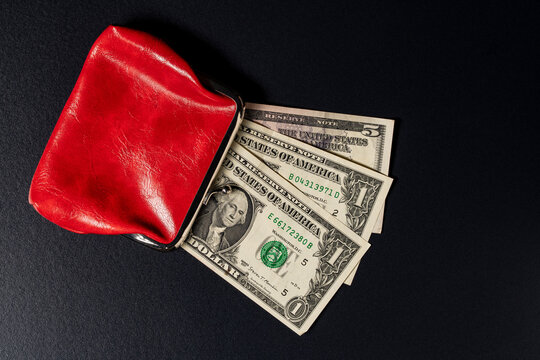 Red leather wallet with US dollar banknotes, top view. Open small red leather vintage clasp purse with one and five US dollar bills peeking out lying on black textured background with copy space