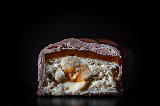 Chocolate bar cross section with nuts nougat and caramel. Closeup shot of cut milk chocolate candy bar showing layers of sweet caramel soft white nougat and crunchy whole hazelnut on black background