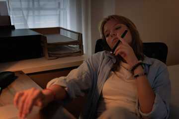 Fototapeta premium Young female student feeling bored and tired during late night study, procrastinating on homework or school project in a dimly lit home office